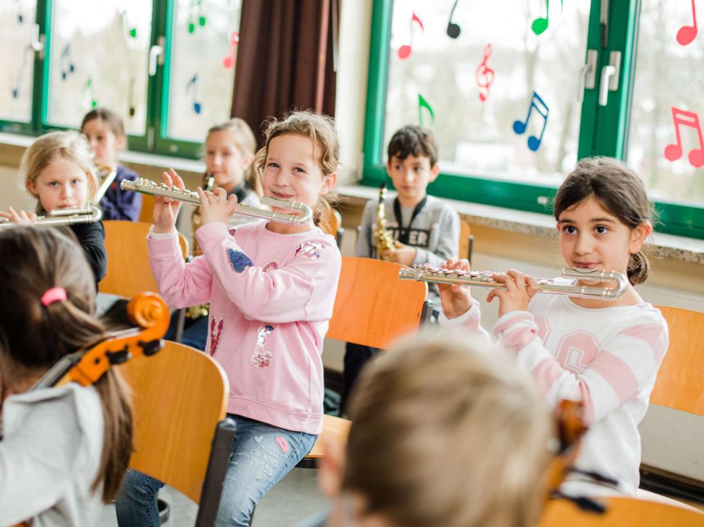 Drei Mädchen spielen Querflöte, davor Cellisten, dahinter Saxophonisten im Klassenraum