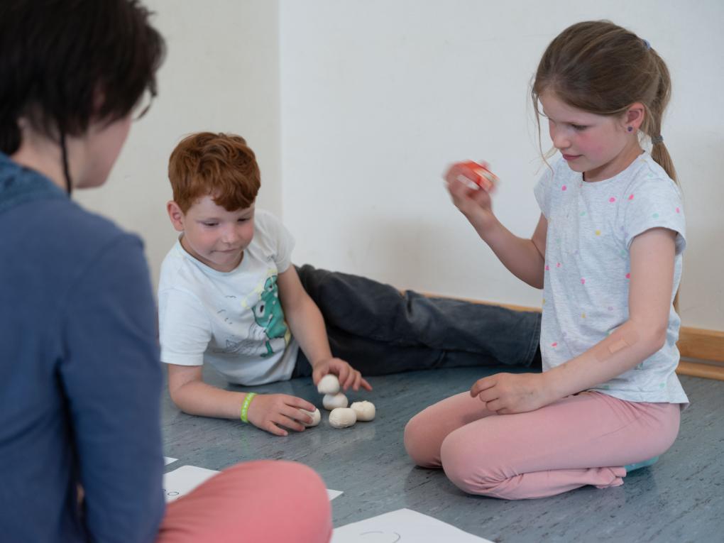 Zwei Kinder spielen auf rasselnden Gegenständen, eine Frau sitzt dabei.